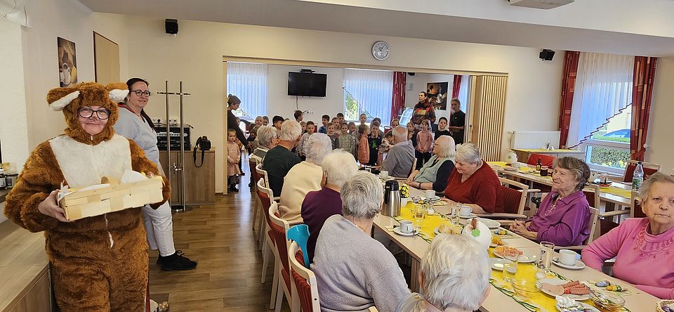 Eine Tafel an der Senioren sitzen, dahinter eine Gruppe Kinder. Vorne links eine Mitarbeiterin im Osterhasenkostüm.