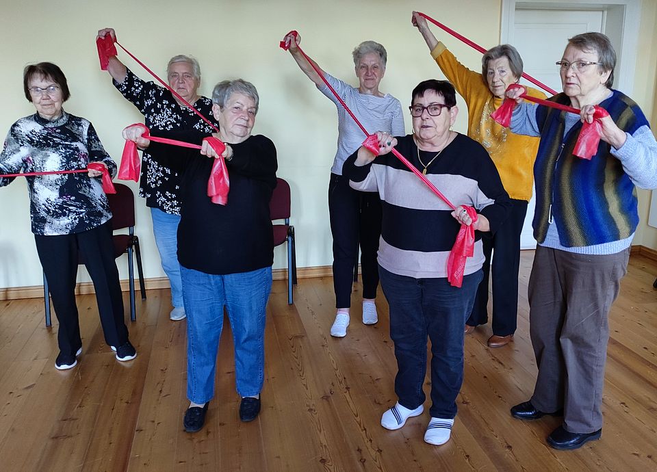 sieben Seniorinnen stehen und dehnen mit Therabändern