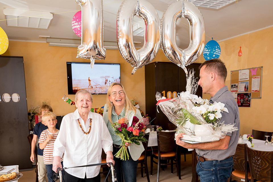 Blumenübergabe an Jubilärin durch Gratulanten. Luftballons in Form einer 100