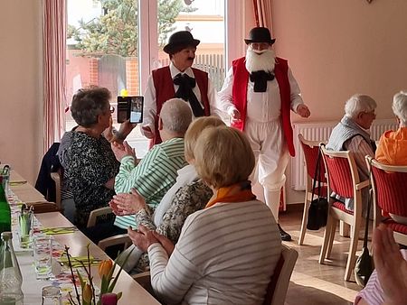 Ein Wildecker Herzbuben Double steh vor einem Fenster. Senioren an einer Tafel klatschen.