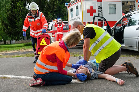 Zwei Ersthelfer in Warnwesten hocken neben einer Verunfallten Person und reanimieren mit Herzdruckmassage (Nachgestellte Szene)