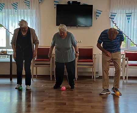 Die Gäste der Tagespflege vergnügten sich beim Kartoffelfest mit Spielen rund um die Knolle. 3 Senioren stehen nebeneinander und schaue herunter zu einem Ball vor ihren Füßen. EIne Kartoffel an einem Band ist um ihre Hüftengebunden und wird als Pendel genutzt.