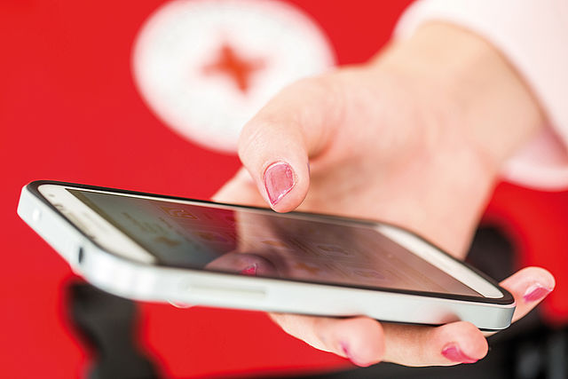 Handy in einer Hand liegend, im Hintergrund das Symbol des Roten Kreuzes