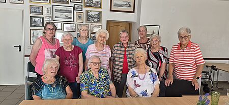 Drei Seniorinnen sitzen an einer Tafel. Dahinter stehen weitere acht Seniorinnen und schauen in die Kamera