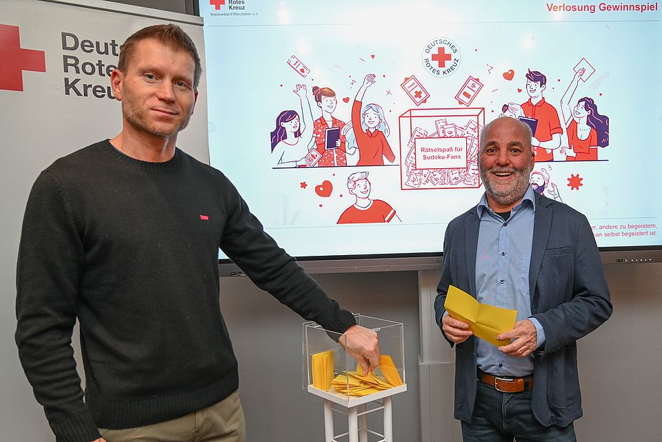 Zwei Männer stehen neben eine gläsernen Würfel mit Losen