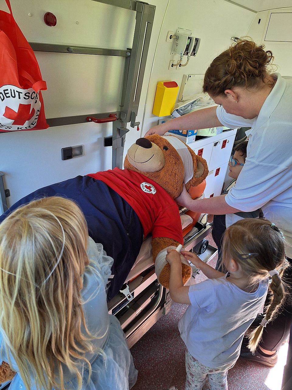 Kinder der Kitasarrezwerge verarzten Bär "Bruno" drei Kindergartenkinder und eine Erwachsen verbinden einen Stoffbären, der auf einer Liege im Rettungswagen liegt