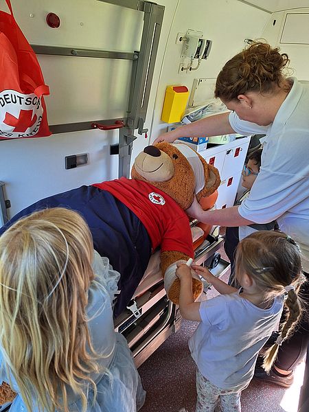 drei Kindergartenkinder und eine Erwachsen verbinden einen Stoffbären, der auf einer Liege im Rettungswagen liegt