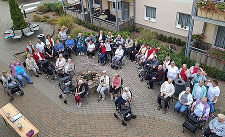Circa 30 Seniorinnen und Senioren, teils in Rollstühlen oder mit Rollatoren winken in die Kamera, die aus dem 2. Stockwerk nach unten fotografiert