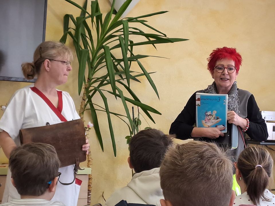 2 Frauen stehen vor einem Publikum und zeigen alte Schulhefte