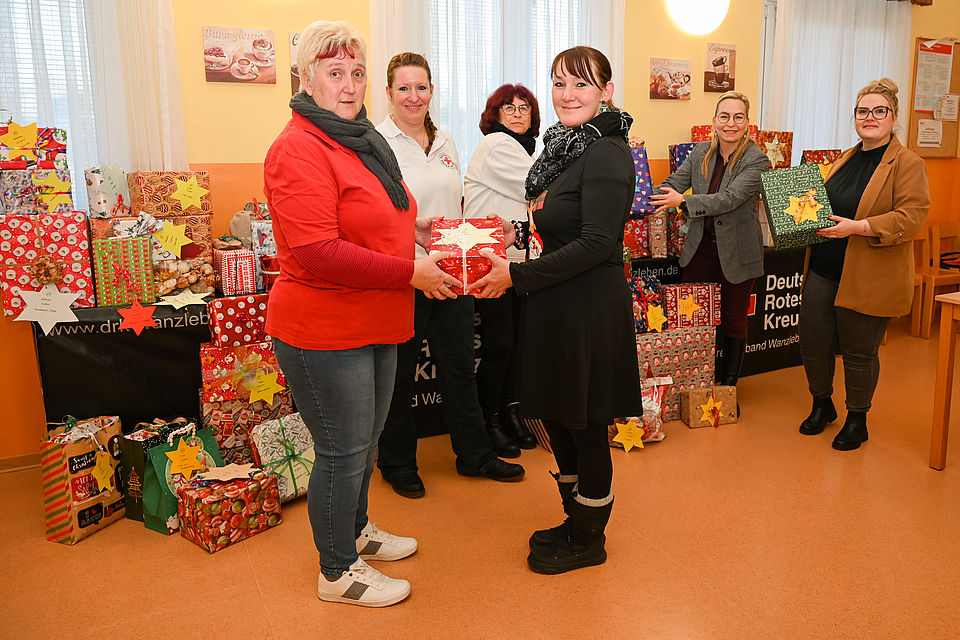 Mehrere Frauen stehen vor einem langen Tisch mit Weihnachtsgeschenke. Eine Frau übergibt einer anderen symbolisch ein Geschenk. 