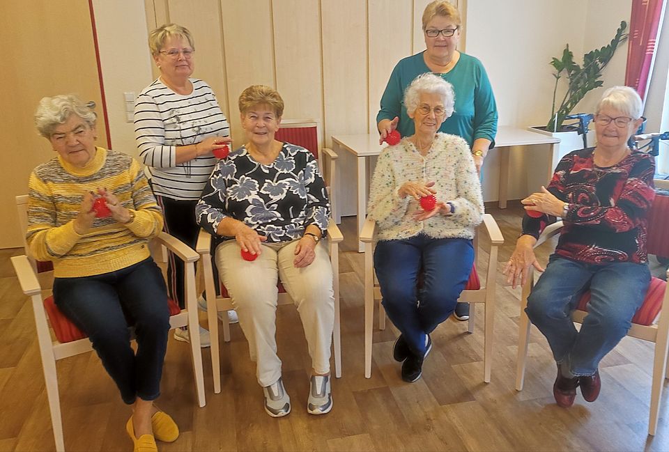 Vier sitzende und zwei stehende Seniorinnen mit roten Igelbällen