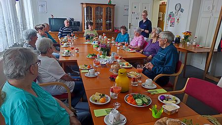 Mehrere Seniorinnen und Senioren sitzen an Tafel mit gesundem Frühstück, wären Apothererin im Stehen etwas erklärt.
