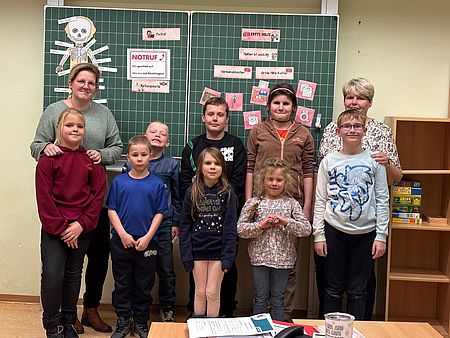 8 Kinder und 2 Erzieherinnen stehen vor einer grünen Tafel mit Bildern zum Thema Erste Hilfe