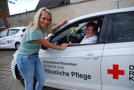 Eine Frau lehnt an einer Autotür. Eine andere Frau sitzt am steuer eines DRK Fahrzeugs