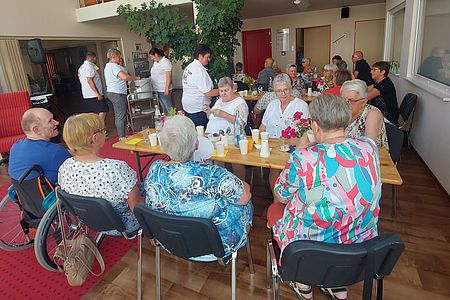 Senioren sitzen an mehreren Tischen. Becher mit Bowle stehen auf den Tischen. Im Hintergrund wir gegrilltes ausgegeben