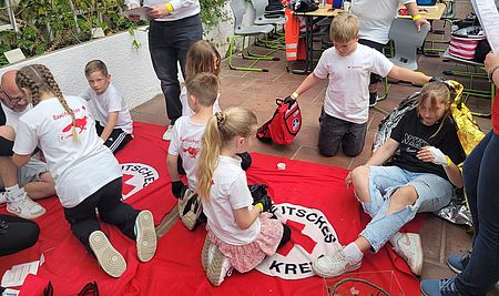mehrere Kinder hocken auf roten Decken und üben die Behandlung von Verletzten