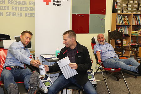 zwei Männer sitzen auf Blutspendestühlen. Einer gibt eine Spende in eine Spendendose