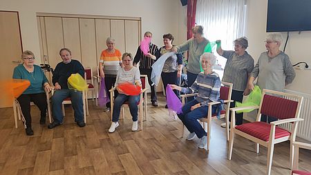 Mehrere Senioren sitzen oder stehen und machen Sport mit bunten Tüchern