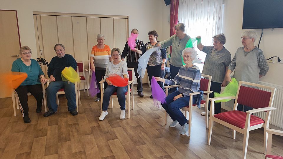 Mehrere Senioren sitzen oder stehen und machen Sport mit bunten Tüchern