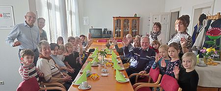 Senioren und Kita-Kinder sitzen an der Geburtstagstafel und winken in die Kamera