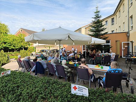 Senioren sitzen unter Sonnenschirmen an zwei Kaffeetafeln im Freien