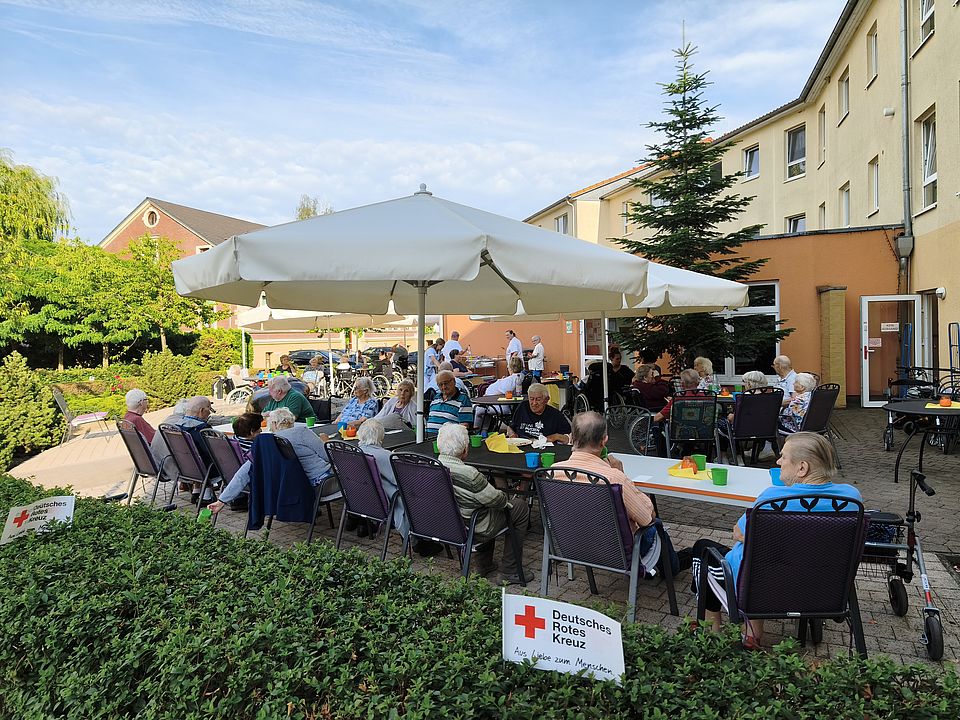 Senioren sitzen unter Sonnenschirmen an zwei Kaffeetafeln im Freien