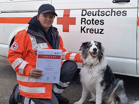 Frau in DRK Einsatzkleidung hockt neben einem Hund und hält eine Urkunde in der Hand