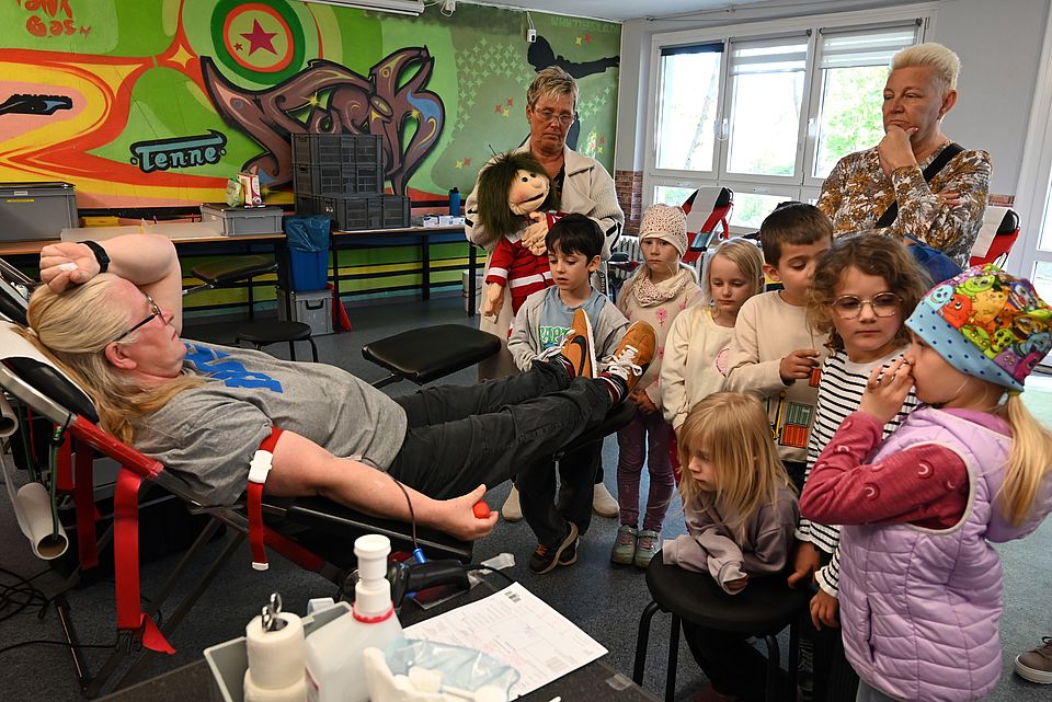Mehrere Kinder und zwei Hortnerinnen stehen neben einer Liege, auf der eine Person liegt und Blutspendet