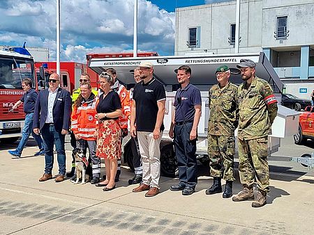 Mehrere Personen, unter Anderem 2 Bundeswehrsoldaten und zwei Sanitäter stehen vor einem großen Anhänger für Hunde
