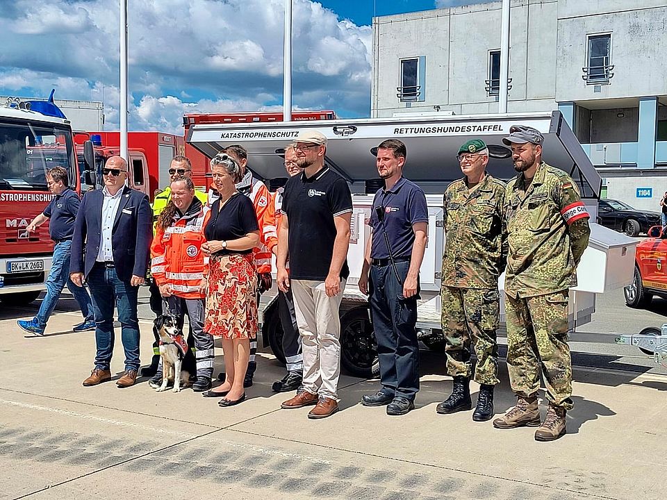 Mehrere Personen, unter Anderem 2 Bundeswehrsoldaten und zwei Sanitäter stehen vor einem großen Anhänger für Hunde